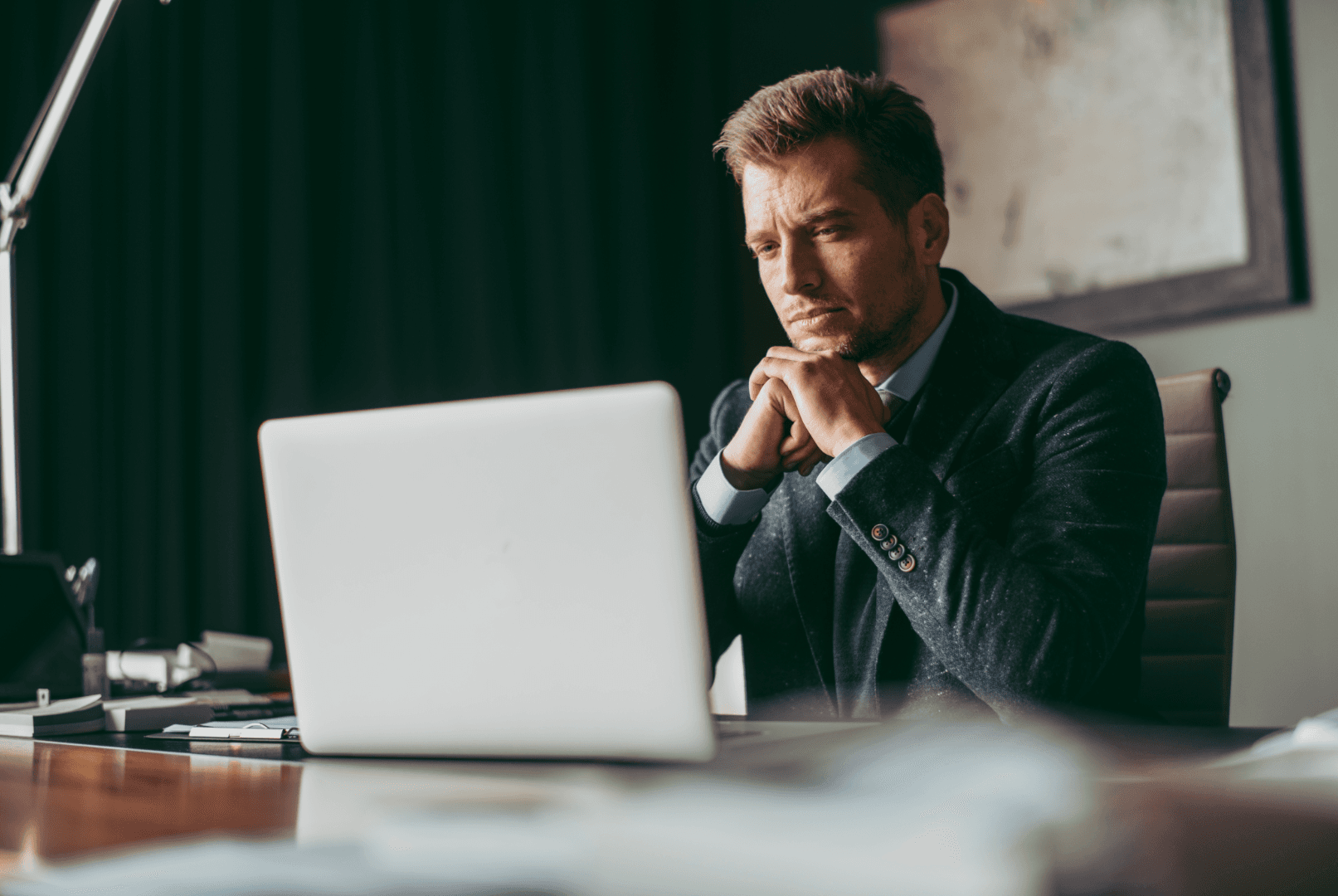 A businessman in a suit sits at his desk, focused on his laptop screen while working
