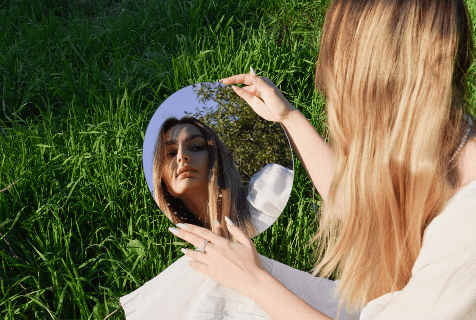 A young woman gazes into a mirror while standing in front of a wide grass field