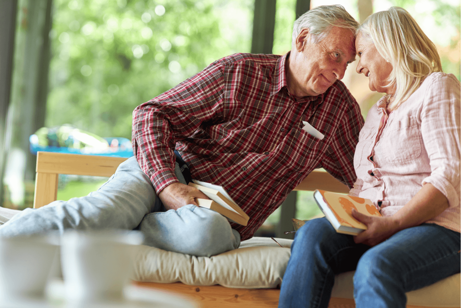 An older man and woman hold books in their hands, smiling gently as they touch foreheads and look into each other’s eyes