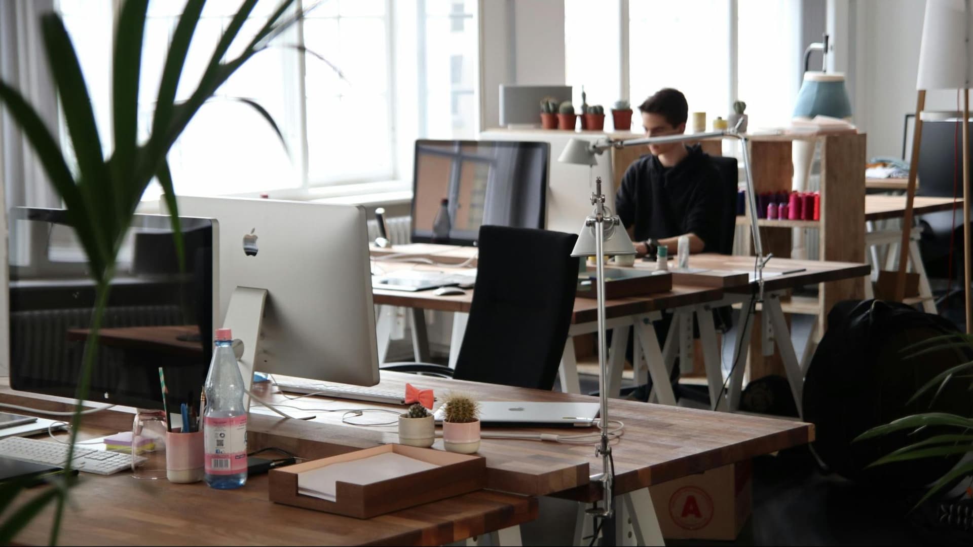 An open-concept office with an empty desk in the foreground and a man working on a computer in the background