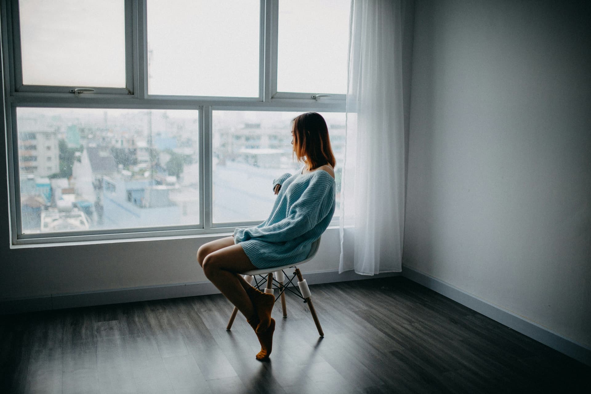 A woman sitting on a chair alone in an empty apartment, looking out the window thoughtfully