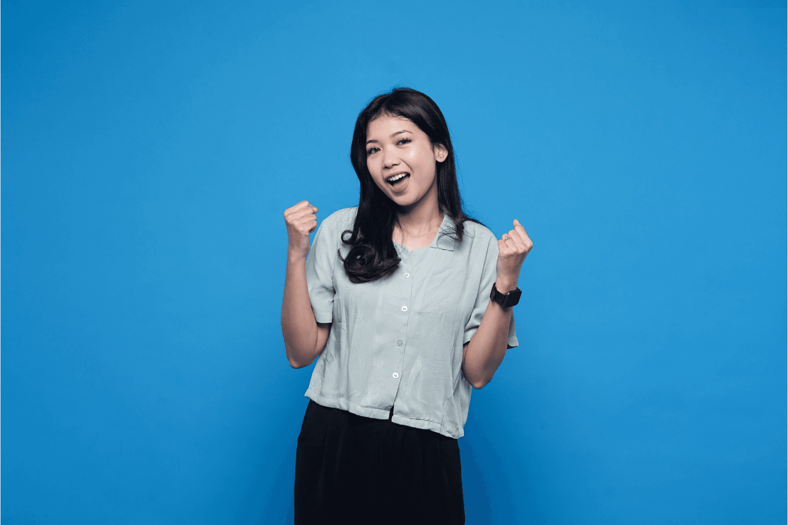 A cheerful woman stands in front of a blue background, laughing and raising her hands in an enthusiastic pose
