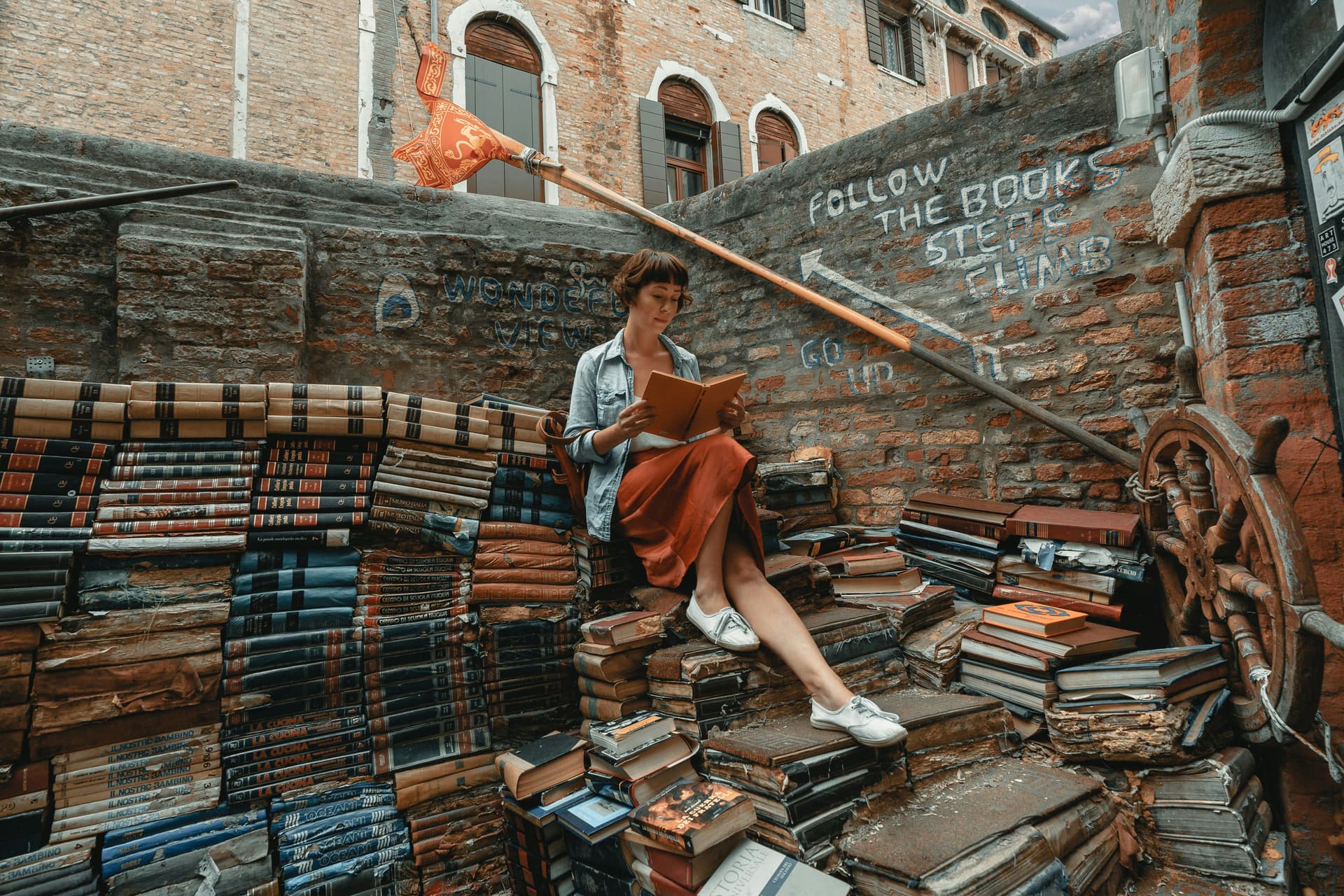 A woman sits on a stairway constructed from stacked books and reads a book