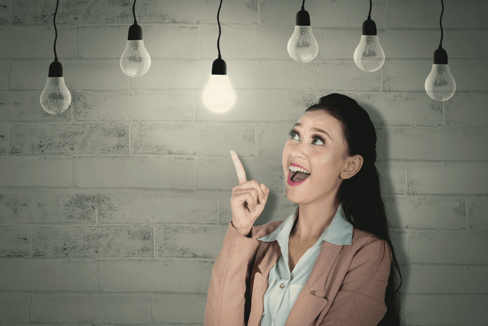 A woman enthusiastically points toward a single illuminated lightbulb, standing out among several turned-off bulbs, symbolizing an idea or inspiration