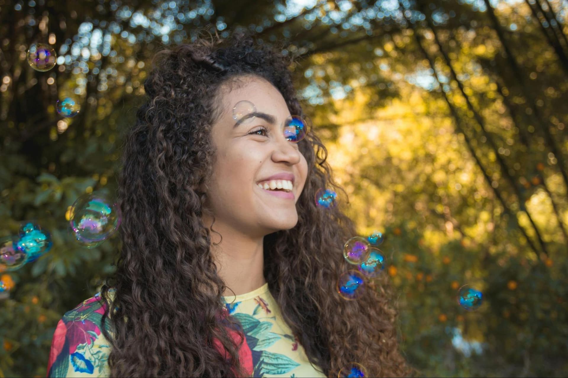 A smiling woman with bubbles flying around her, with trees behind her