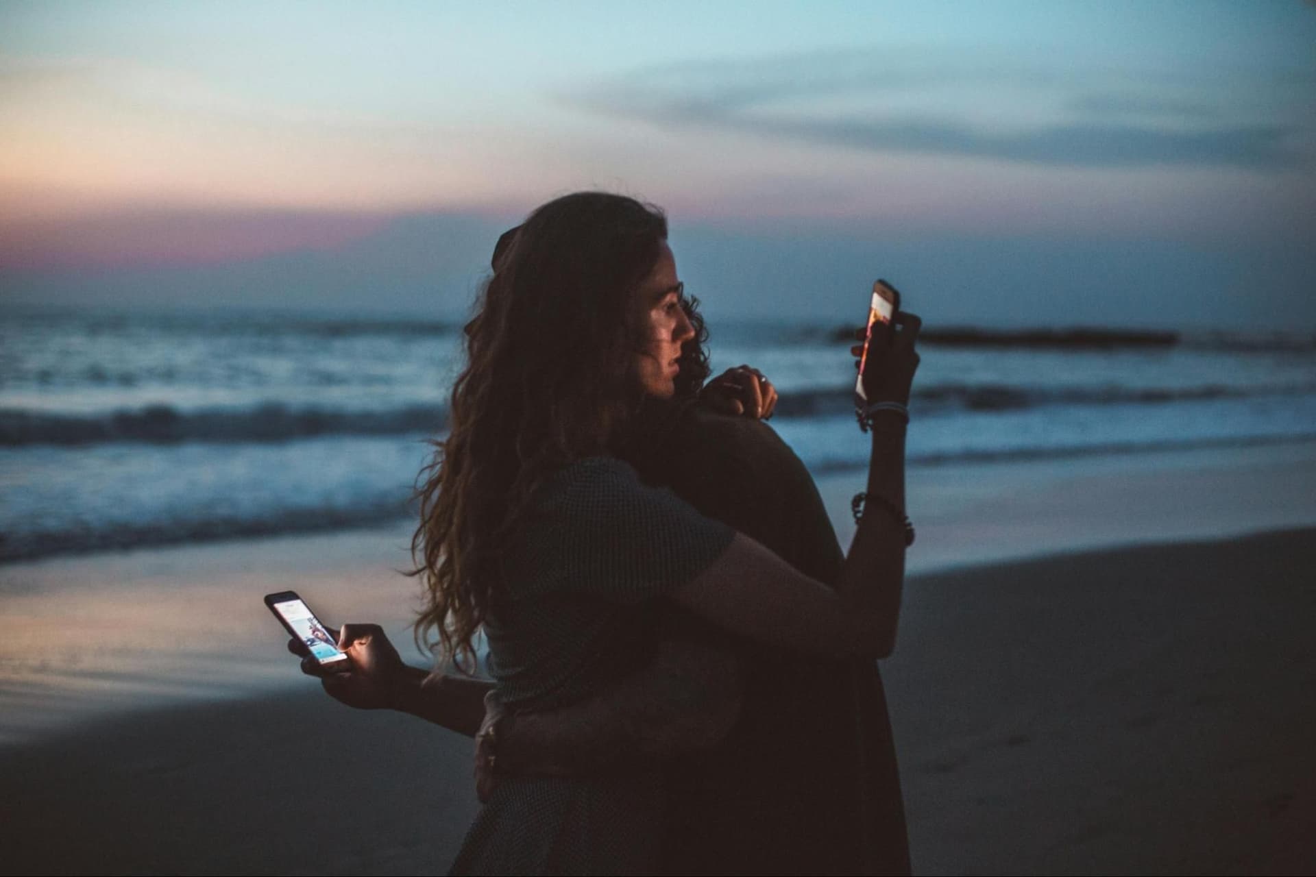 Two people hugging by the ocean while both look at their phones