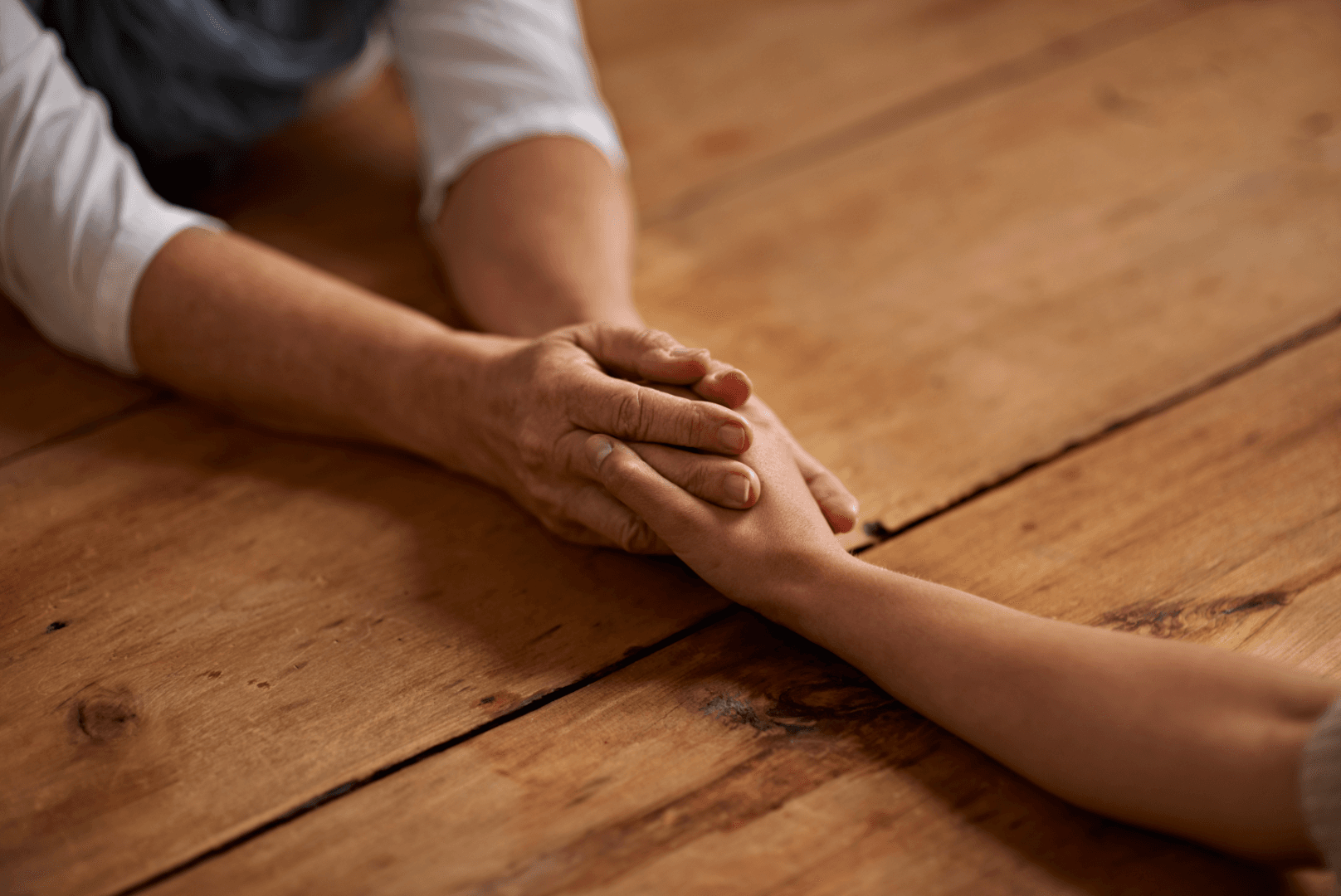 Close-up of two people’s hands holding each other while resting on a wooden floor