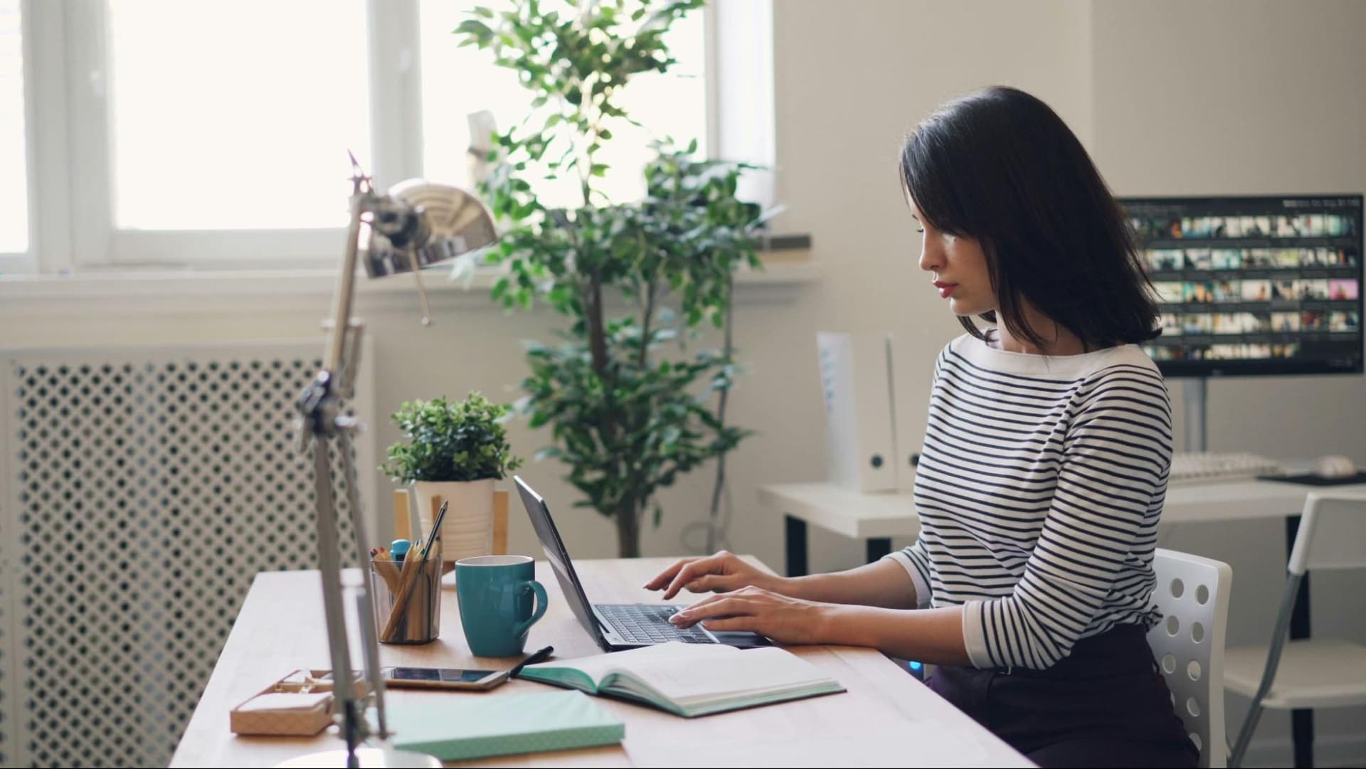 a woman focused on her work at a tidy desk, surrounded by plants and a lamp
