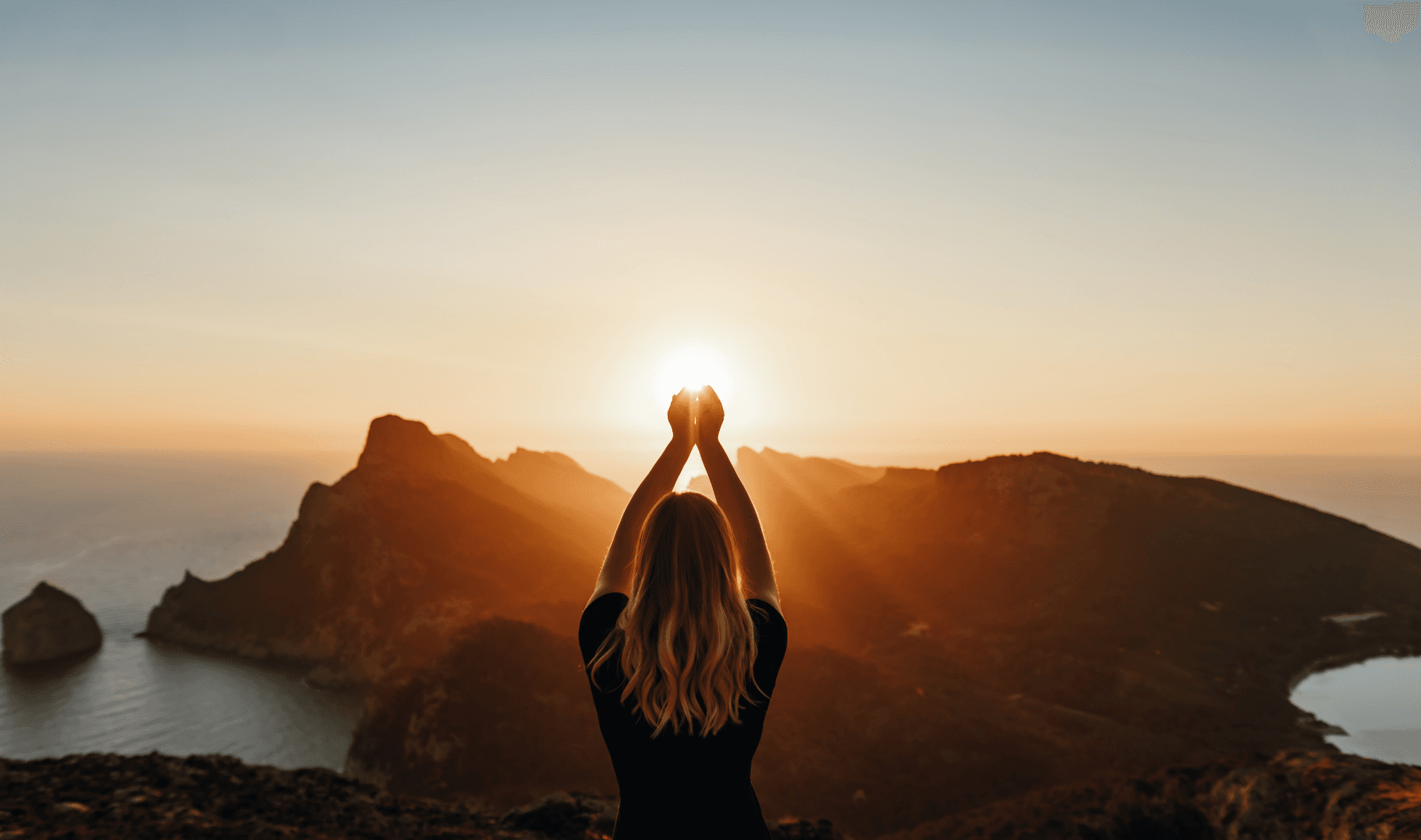 A woman facing a mountain and beach view, reaching her hands toward the sun as if holding it