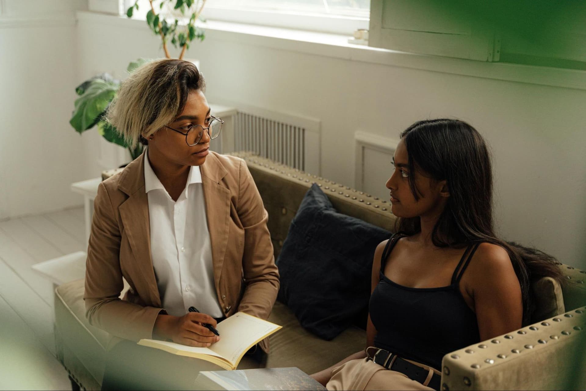 Two women sitting on a couch talking, with one writing in a notebook