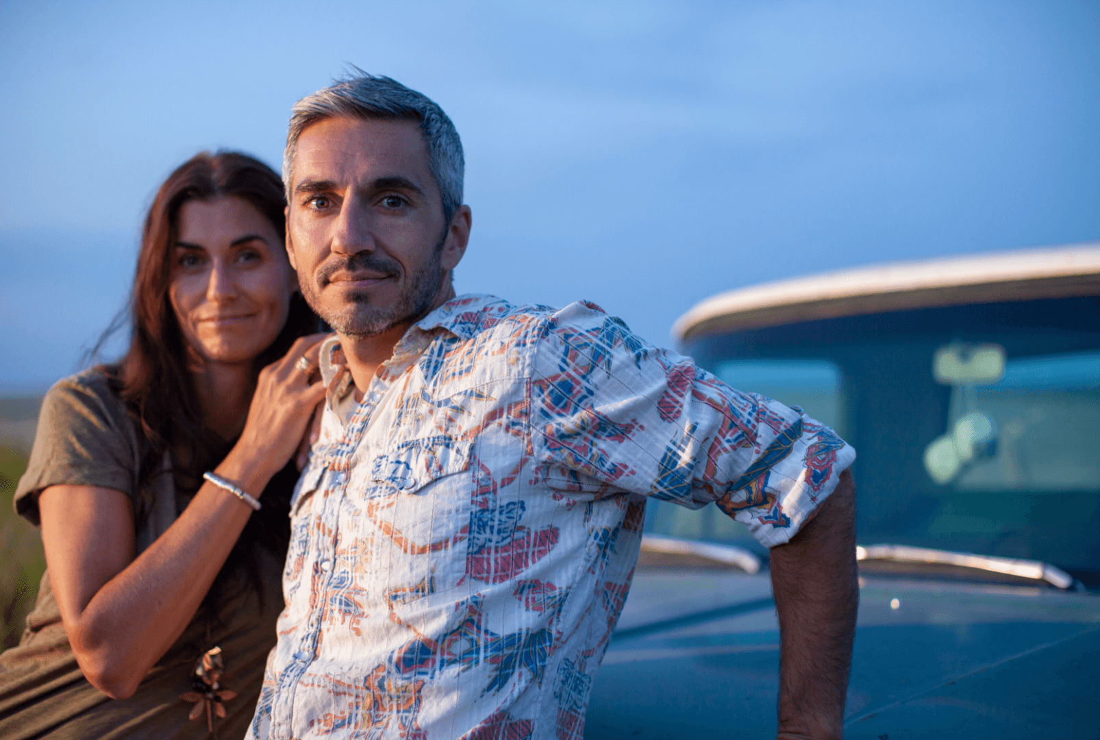 A man and a woman looking at the camera, with the woman resting her arms on the man’s shoulder