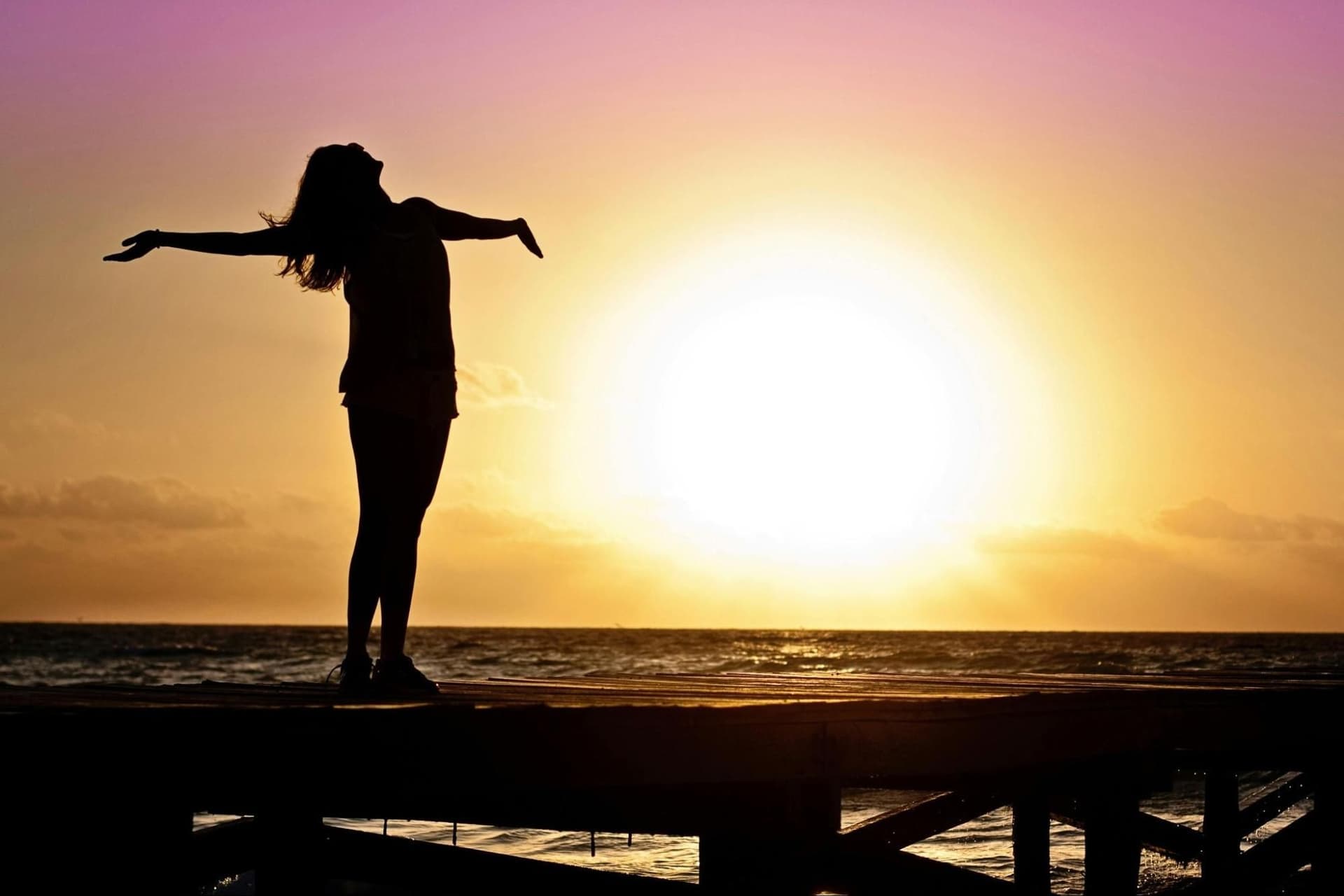 A woman’s silhouette stands on a beach in dim light, arms outstretched as if soaking in the sun