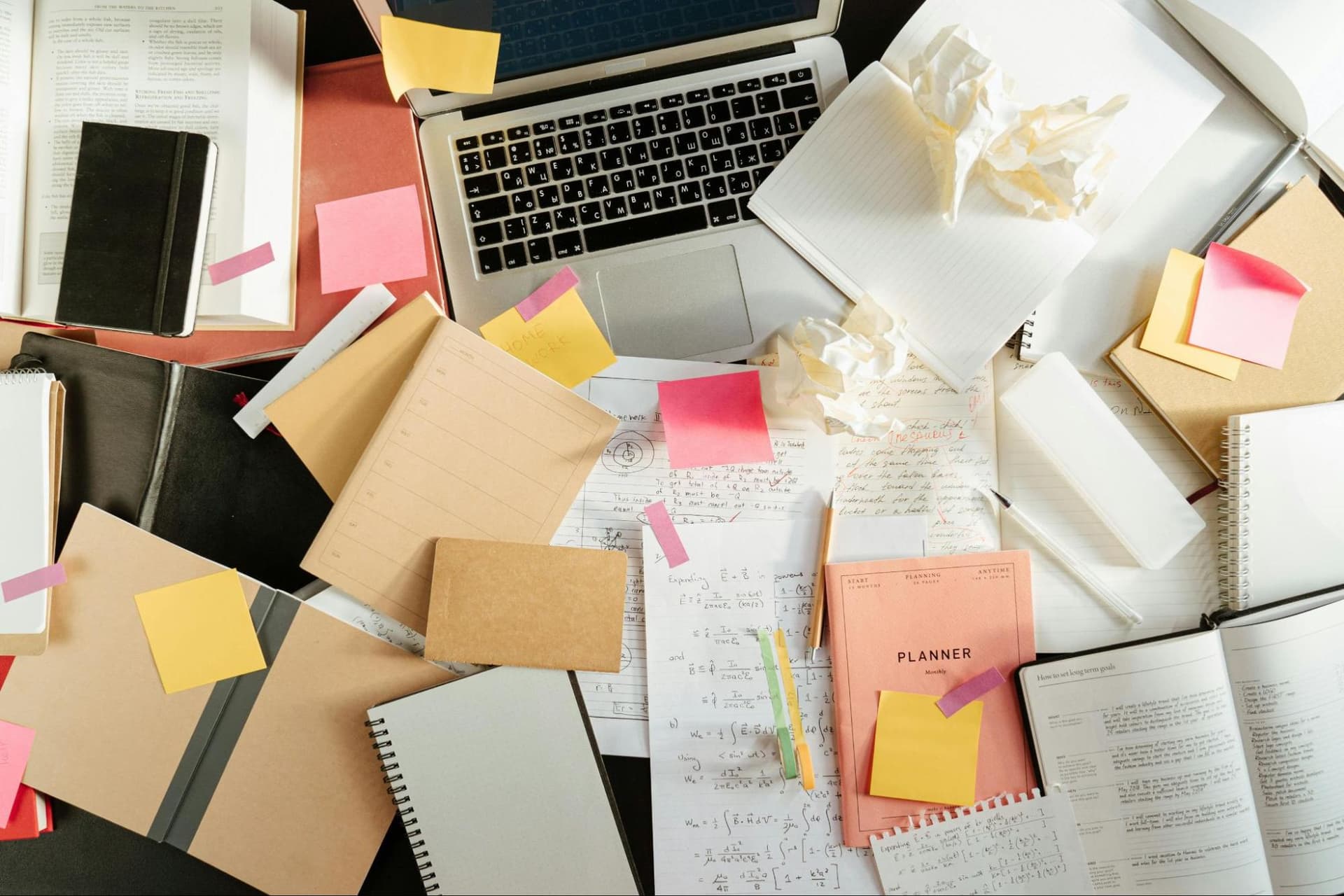 A messy table covered with notebooks and papers, with an open laptop among them