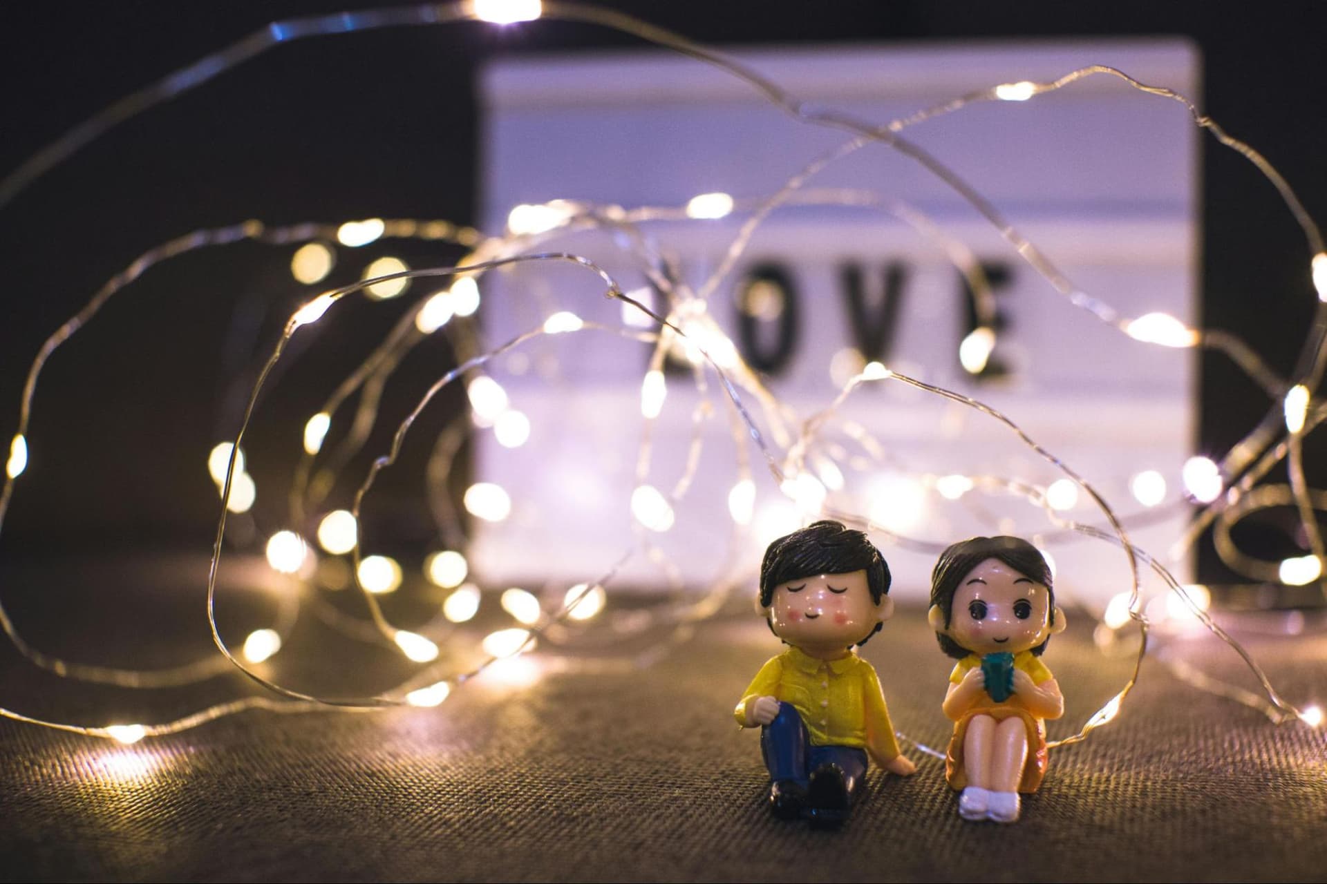 Figurines of a man and a woman surrounded by soft decorative lights, with a glowing neon “love” sign behind them