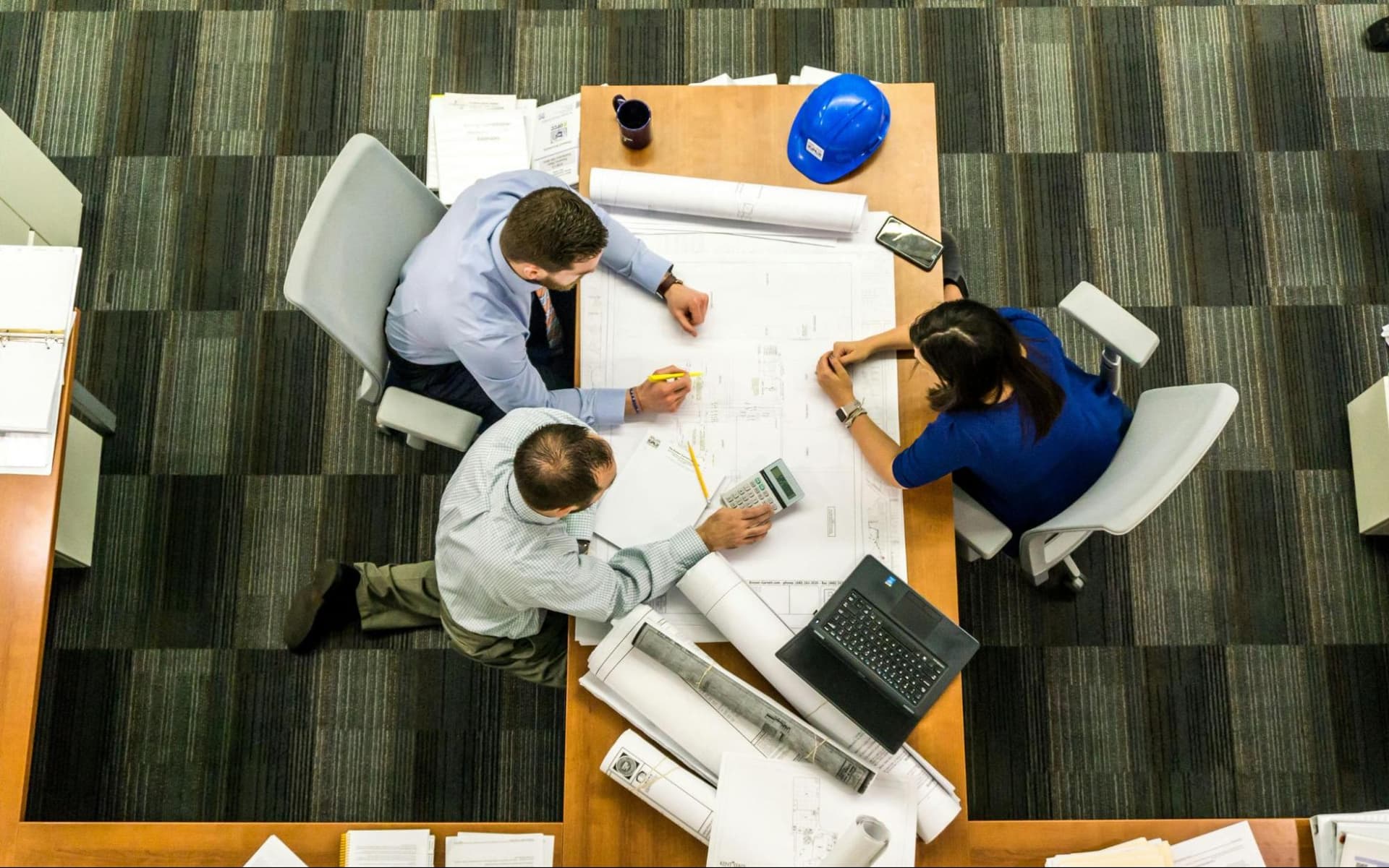 Three colleagues sitting around an office table examining a big paper draft, while one person uses a calculator