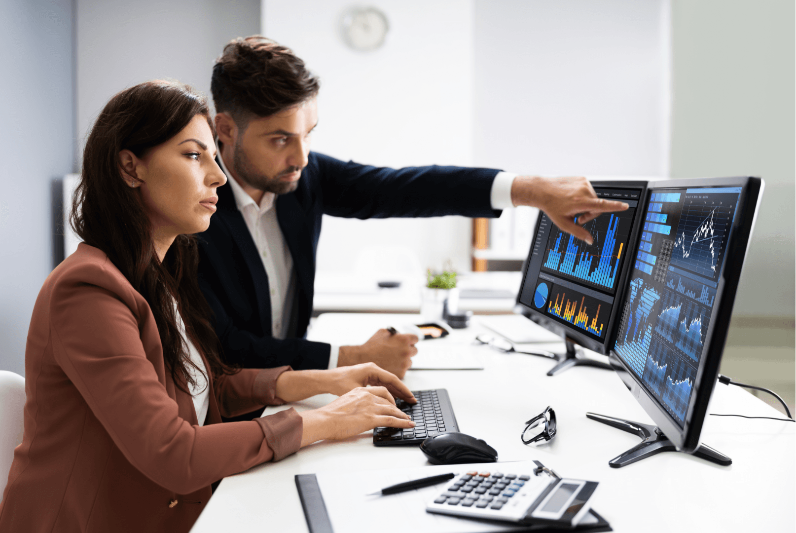 A man and a woman in a workspace look at a computer screen displaying graphs and data