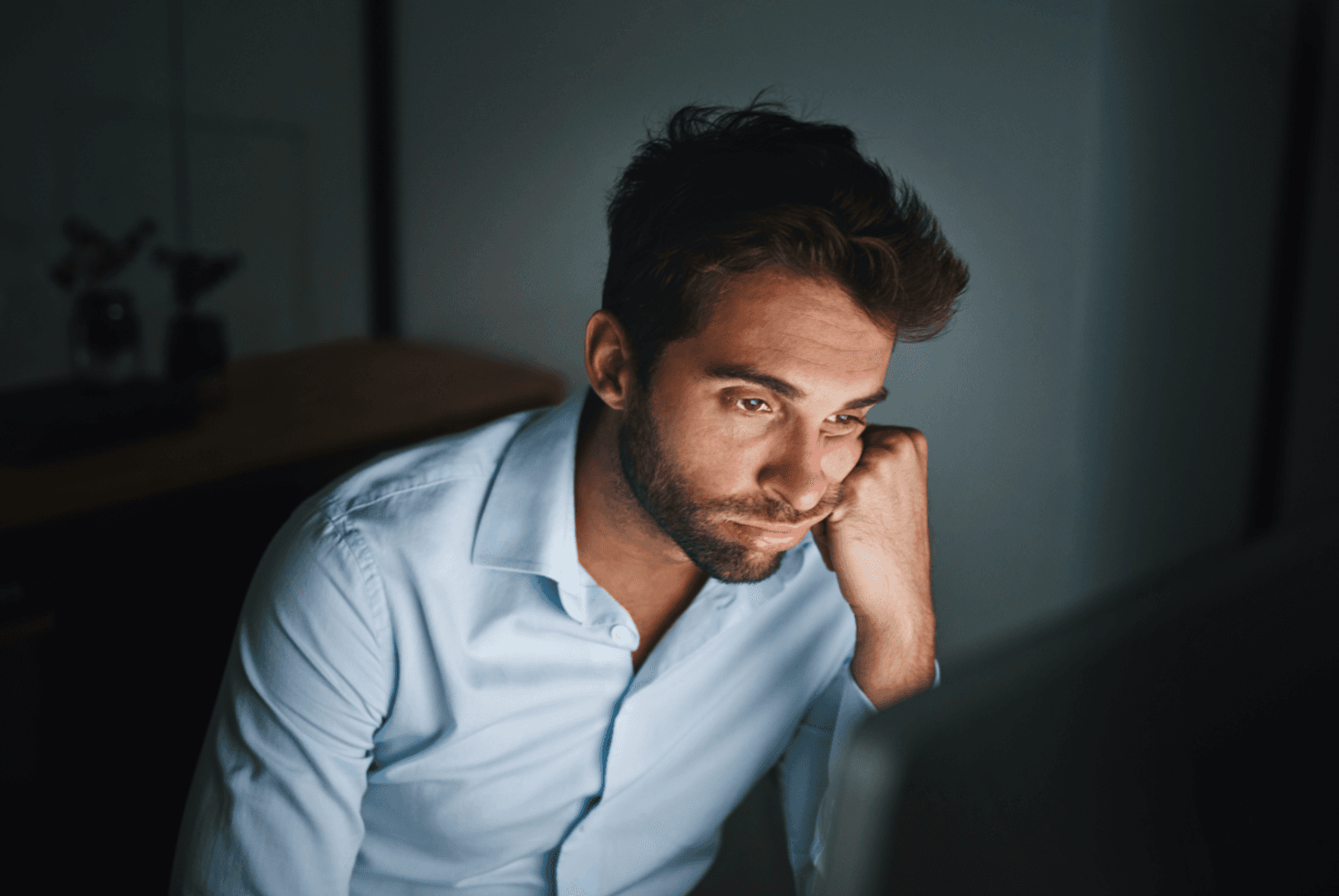 A man sits in dim light, staring at a screen with a bored expression