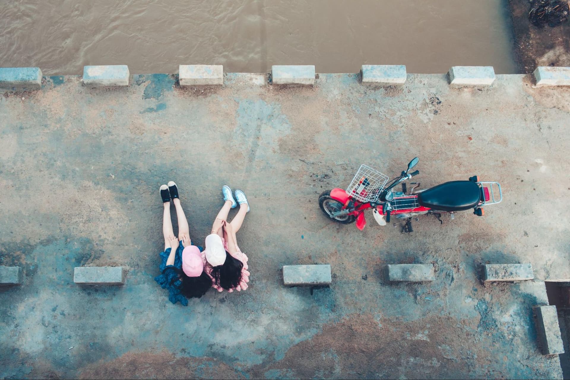 two young women sitting on a concrete river bank with a scooter next to them