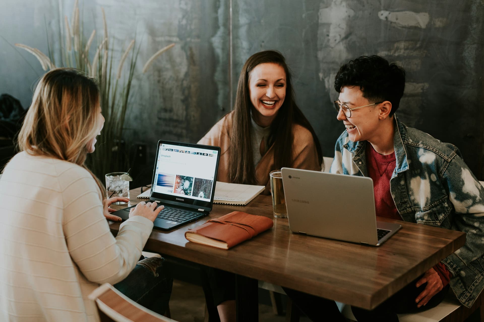 Three people sit around a table with open laptops, smiling and laughing during a conversation