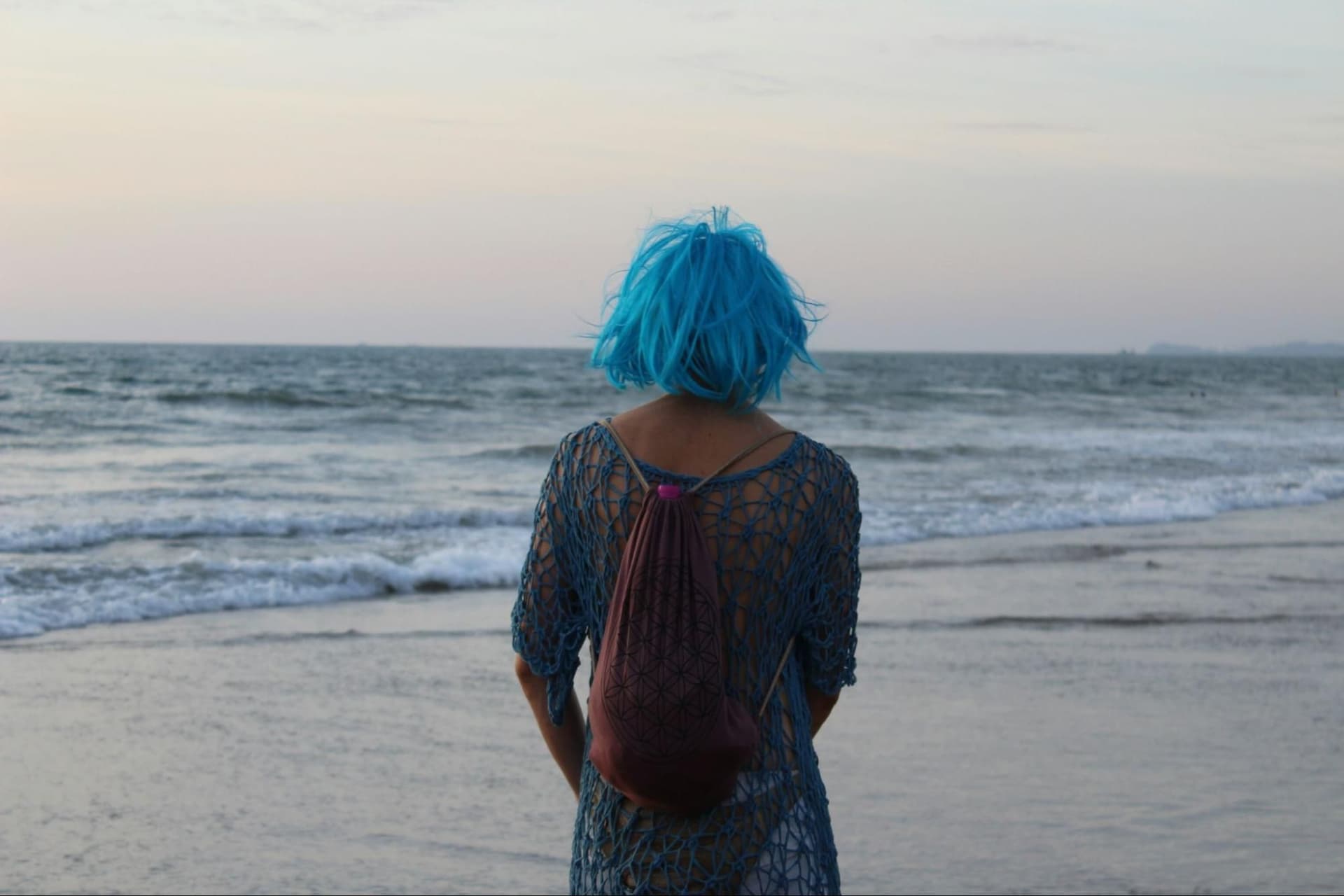 A woman with blue hair stands facing the ocean, gazing out at the water
