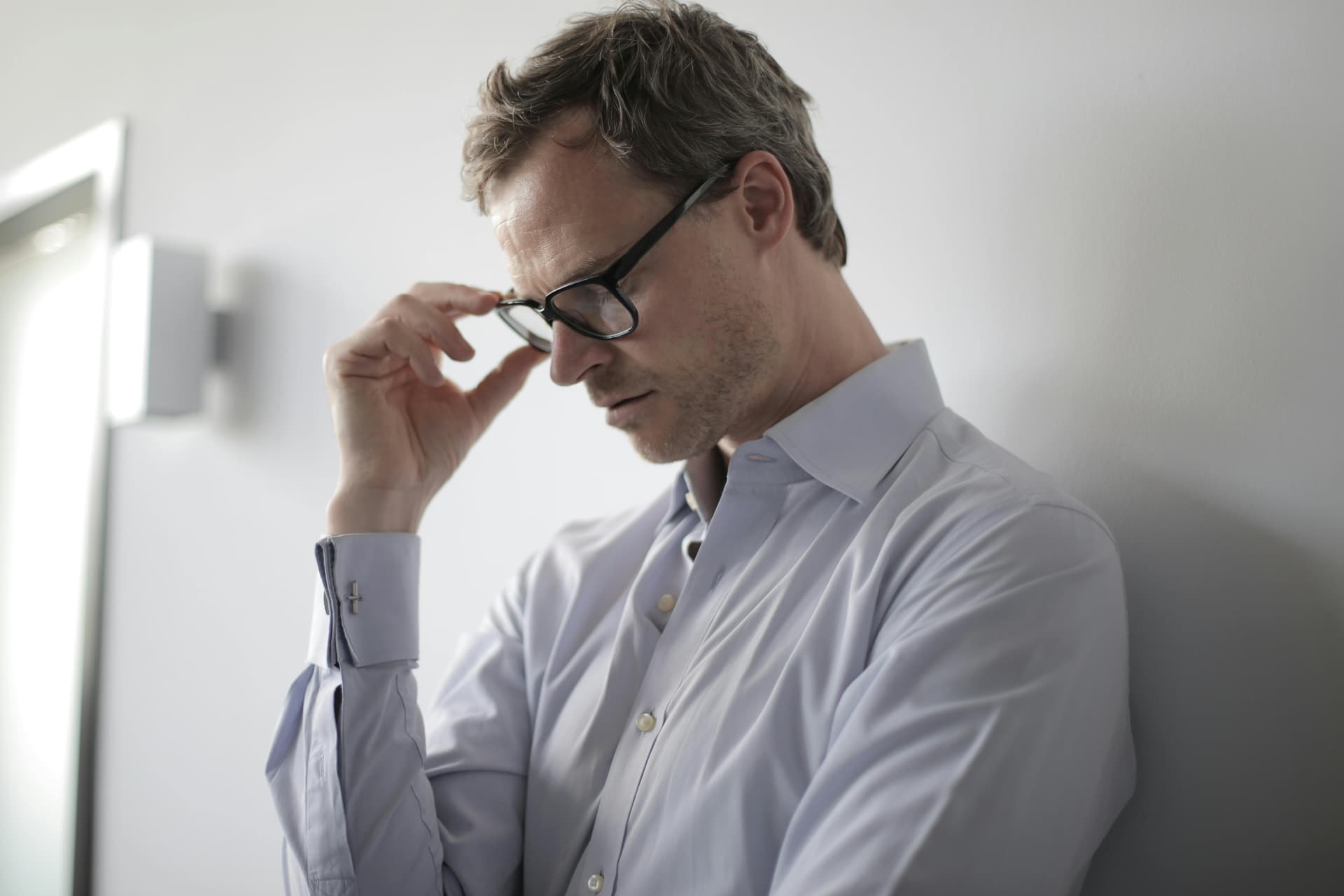 A man wearing black eyeglasses holds his head and looks down, showing a clear expression of stress and emotional strain