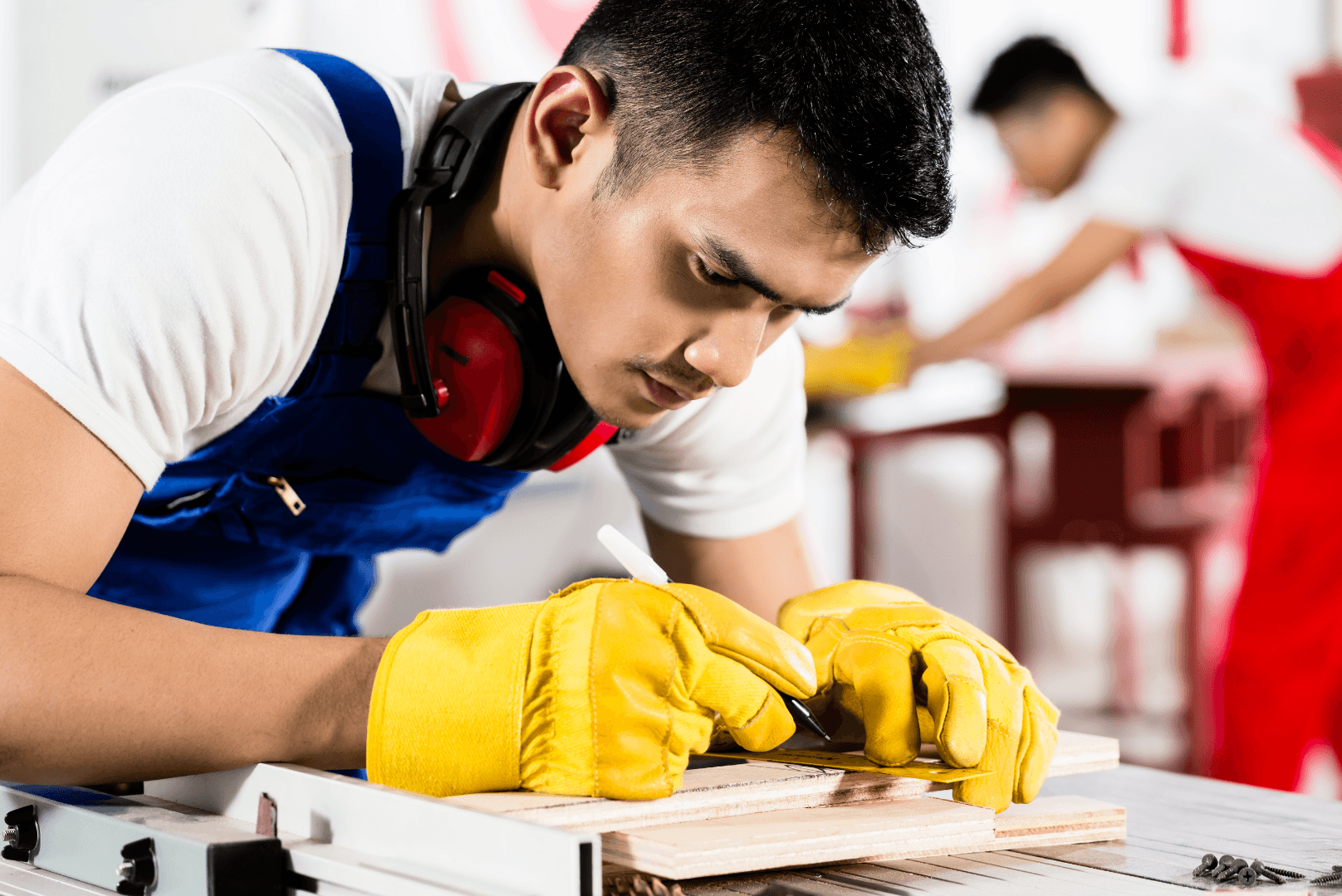 A man wearing a work uniform uses a measuring tape and pencil to mark measurements on a piece of wood