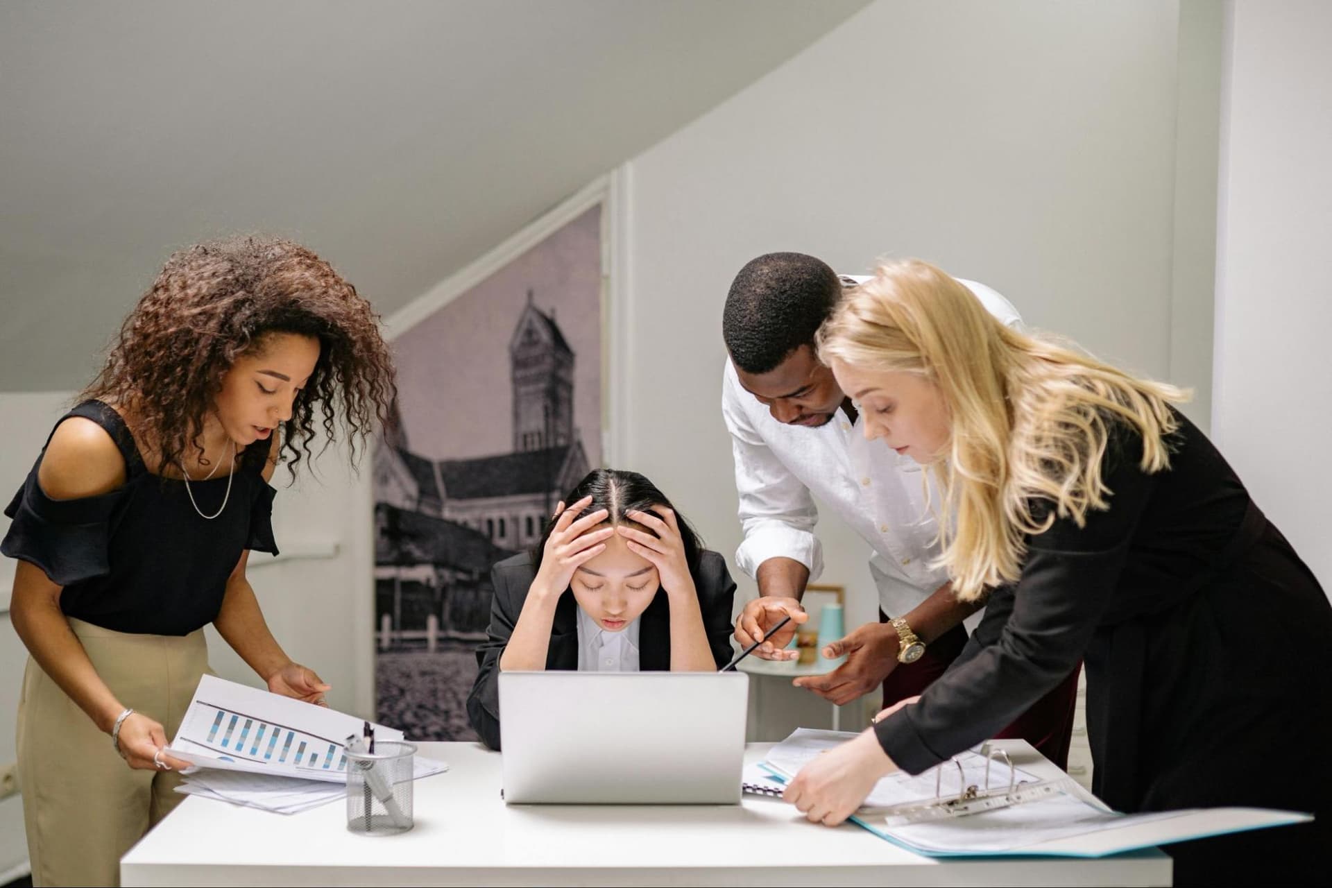A stressed woman sits at her desk with a laptop, holding her head in her hands as three colleagues stand nearby looking at documents, showing a busy work environment